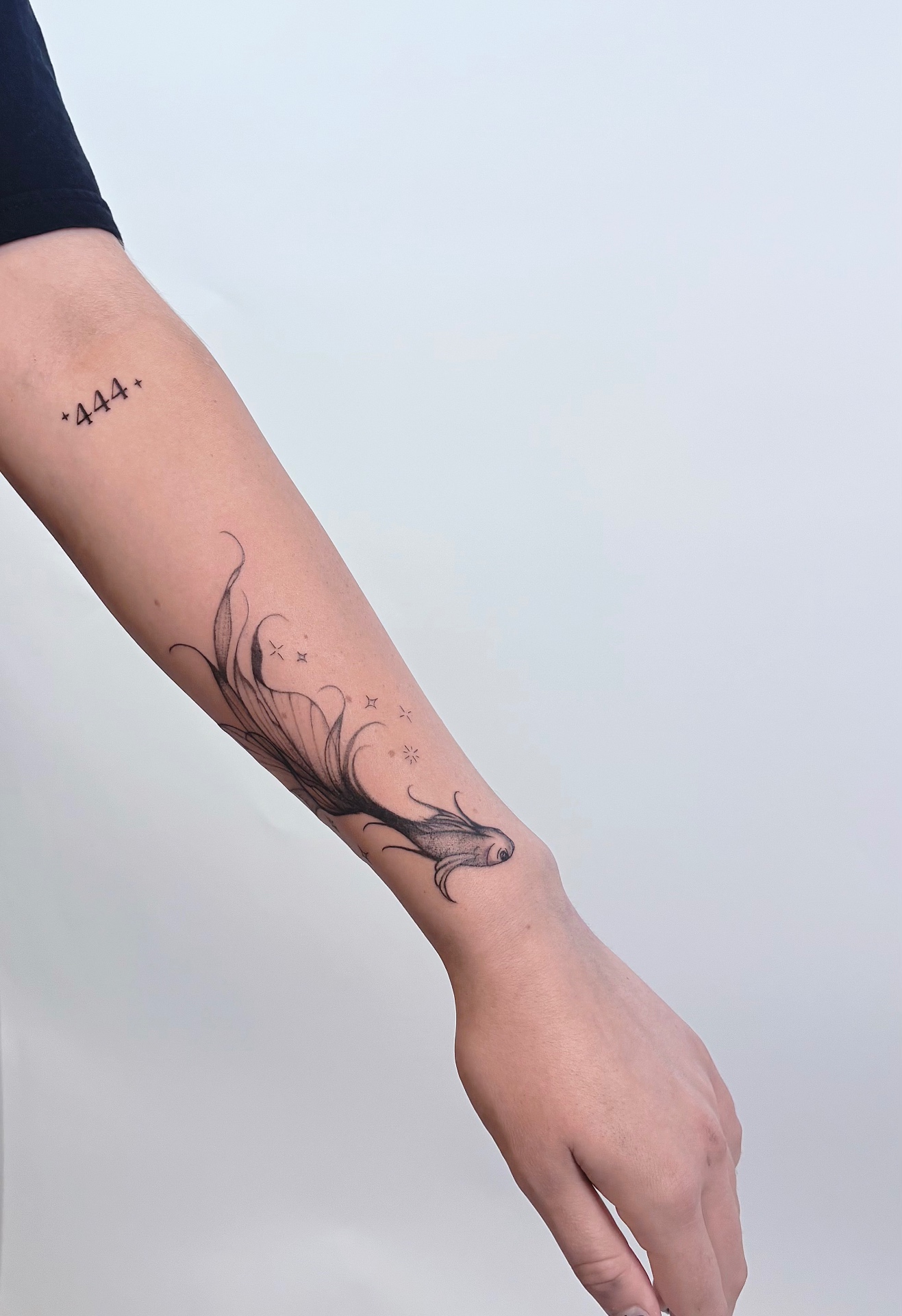 Fine line fish with flowing fins and small sparkles tattooed on an inner forearm