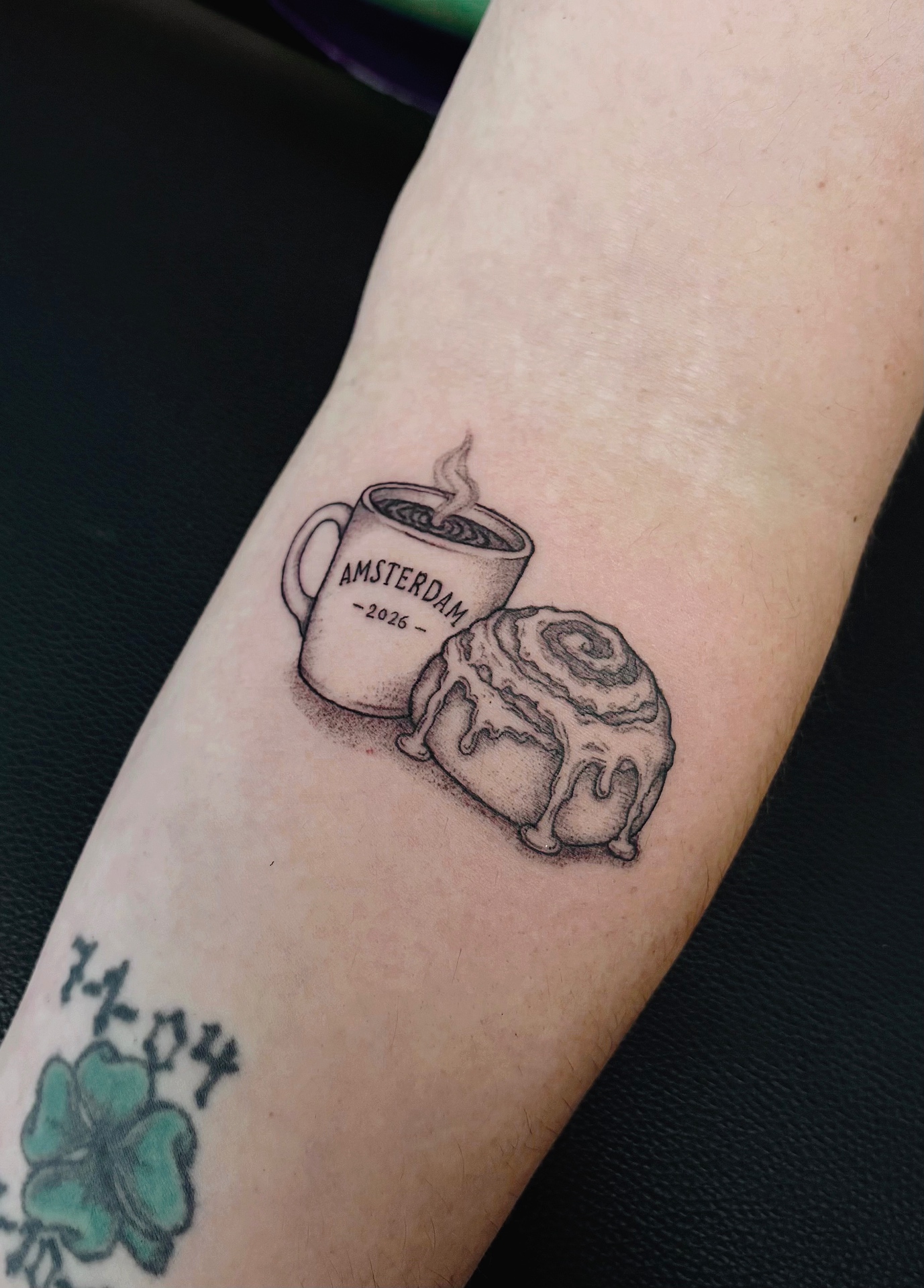 Coffee mug marked Amsterdam 2026 beside a cinnamon roll in fine dotwork on an inner forearm