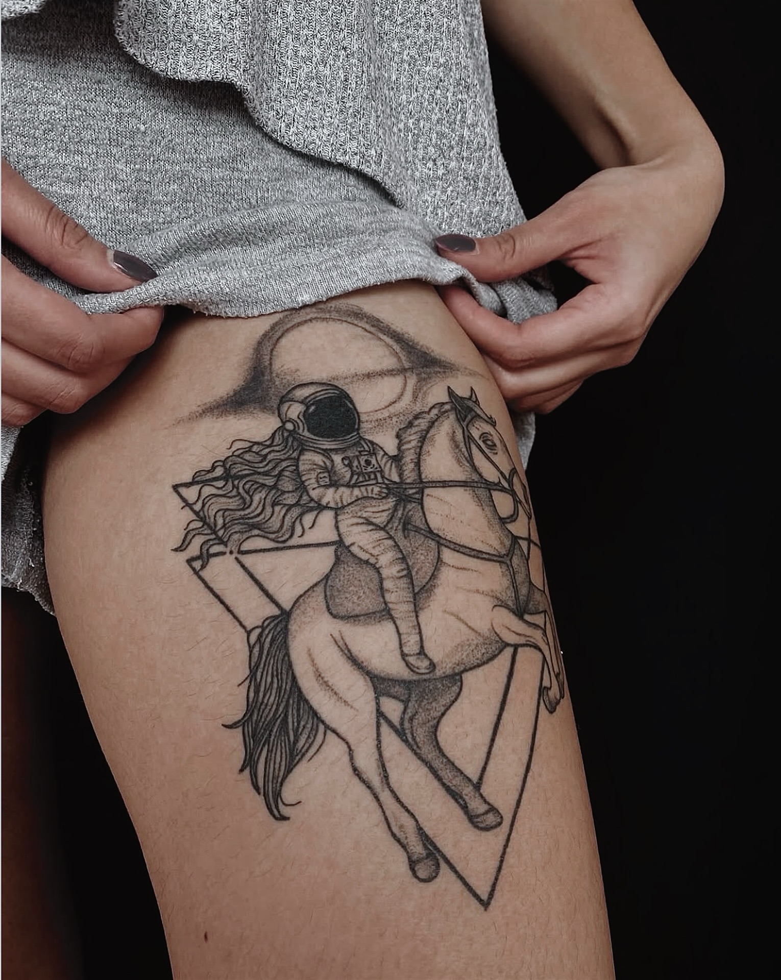 Astronaut with long hair riding a horse before a ringed planet inside framed triangles, in dotwork on a thigh