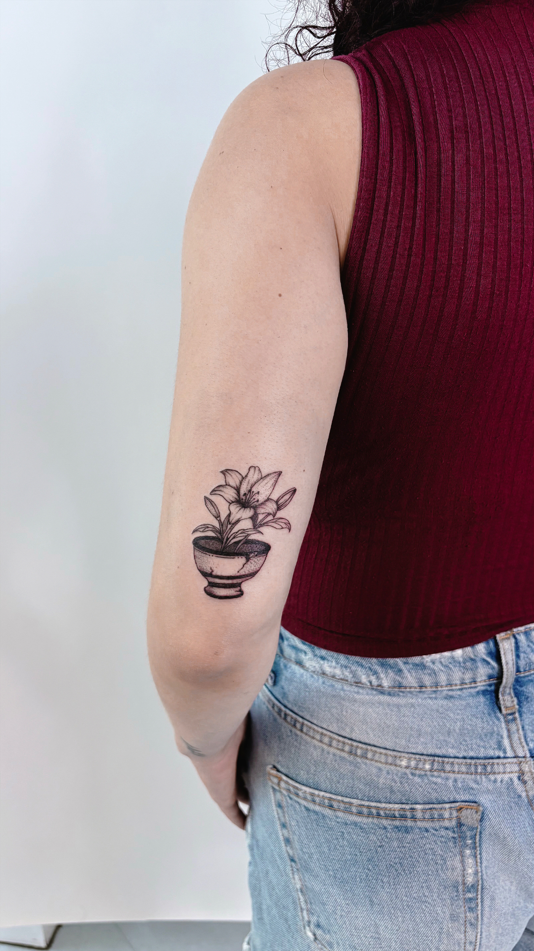 Lily growing from a small ceramic pot in fine line dotwork above the elbow